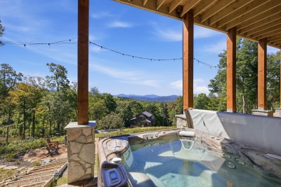 Soak in the private hot tub with stunning mountain views stretching past the treetops. It seats eight on the covered lower patio, string lights glow overhead after dark, and the fire pit circle waits just down the timber steps for the next round.