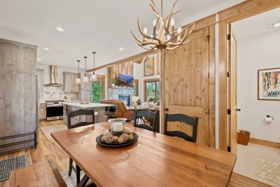 Rustic open-concept dining, kitchen, and fireplace living area, all in one easy sightline. Pass plates from the island to the farmhouse table under the antler chandelier while the stone fireplace glows across the room and the half bath sits nearby.