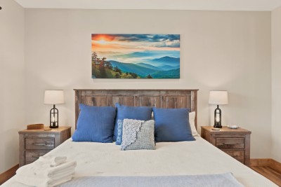 Primary king bedroom with mountain-inspired decor, from the sunrise-over-the-ridges canvas to the soft blue pillows. Weathered wood nightstands and lantern lamps frame the bed, and fresh towels are folded and waiting at the foot when you arrive.