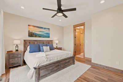 Main-level primary bedroom with king bed, wood accents, and bright, airy comfort. A reclaimed-wood bed frame, matching nightstands, a ceiling fan and blackout shades set up a quiet night, with the private ensuite bath right through the open door.