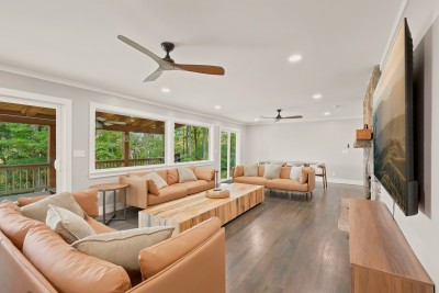 The main living room wraps two leather sectionals around a slab coffee table, with a big smart TV and a propane fireplace set in stone. A wall of windows and a glass door look onto the covered deck and the hardwoods, bringing the woods inside