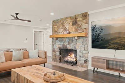 Living room with a tall stacked-stone fireplace with a lit fire and a light wood mantel, a large wall-mounted TV over a wood and glass media console, a tan leather sofa with pillows, a wood coffee table with a woven tray, and a ceiling fan above
