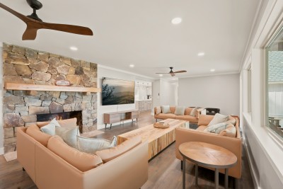 From the window end of the main living room, two leather sectionals face each other across a long slab coffee table, with the stone fireplace at one side and the kitchen in view beyond. Plenty of seats for the whole group to gather after dinner