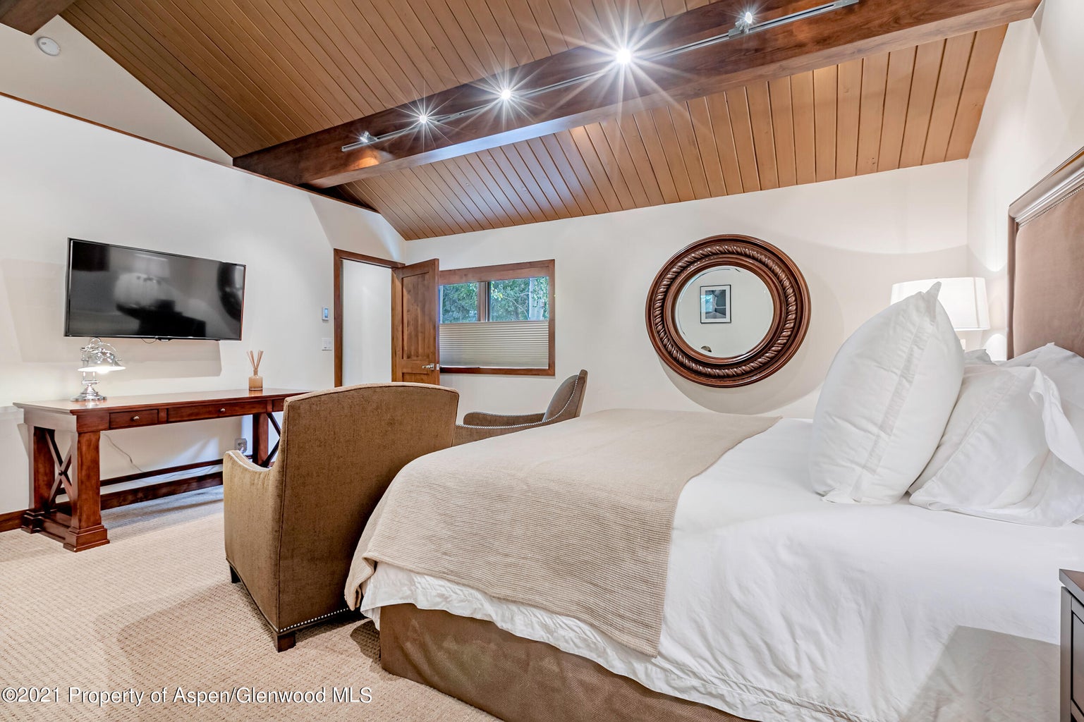 Cozy vaulted bedroom with plush bedding, TV, desk, and warm wood details
