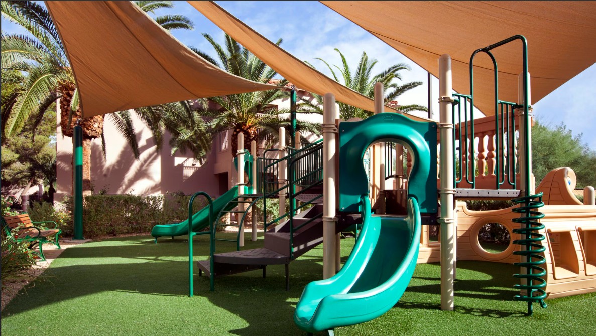 Shaded family playground with slides, soft turf, and tropical surroundings