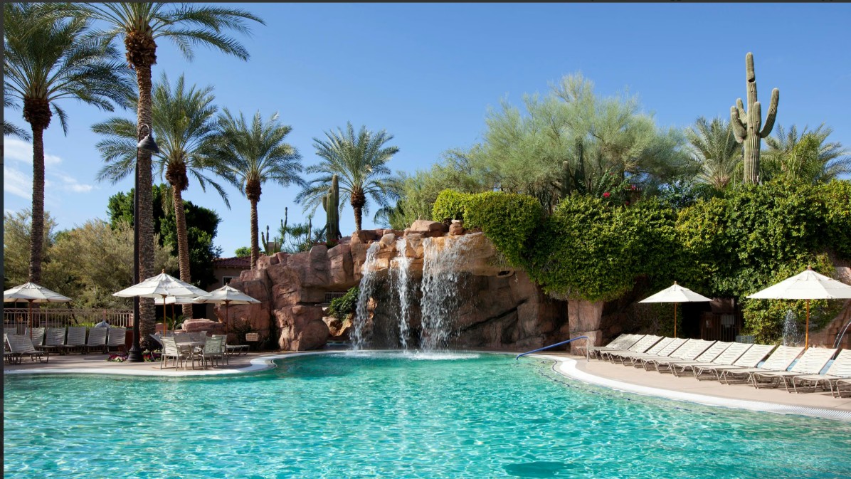 Resort-style pool with waterfall, palms, and sunny lounge seating