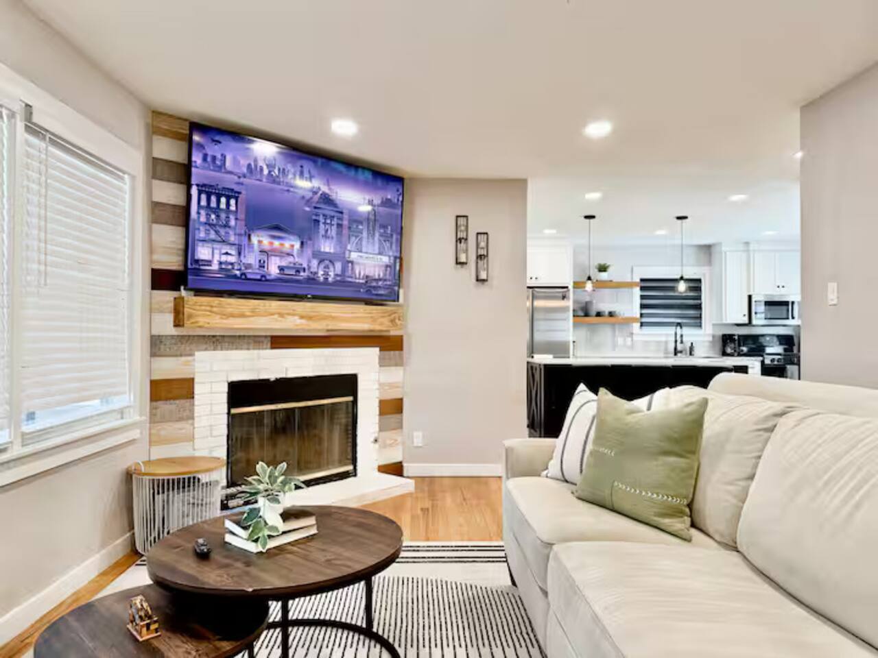 Relax in this modern, light-filled living space featuring a cozy fireplace, rustic wood accents, and a large smart TV. The open-concept layout flows into a sleek kitchen, perfect for a stylish and comfortable getaway.