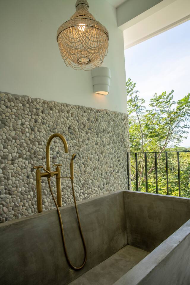 A custom outdoor soaking tub, bathed in chandelier light, opens to sweeping treetop vistas and a serene glimpse of the sea.