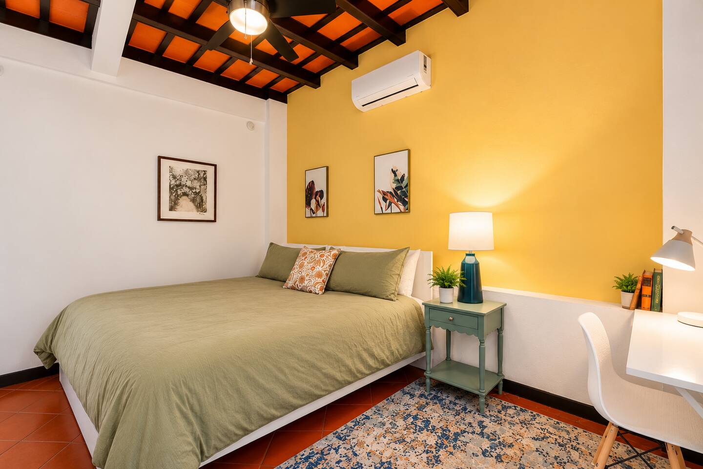 Unwind in this bright, inviting bedroom featuring warm Caribbean colors, exposed wood-beam ceilings, a comfortable bed, quiet air conditioning, and a ceiling fan. A dedicated workspace or makeup station adds convenience to your Old San Juan stay.