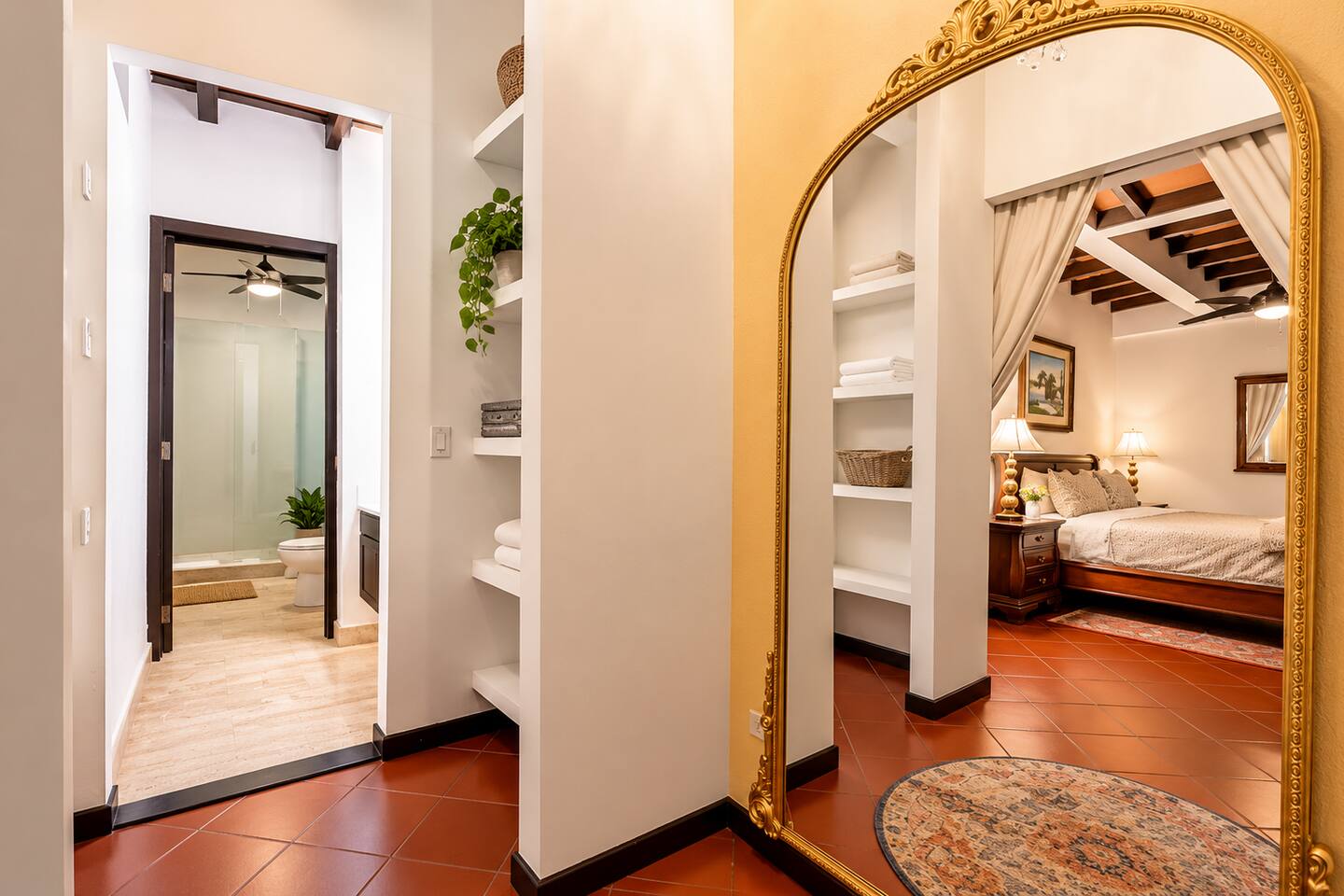 A touch of Old San Juan elegance! This charming dressing area features an ornate gold mirror, built-in shelving, & warm terracotta floors. The adjoining bathroom offers a spacious walk-in shower, while the mirror reflects the inviting primary bedroom