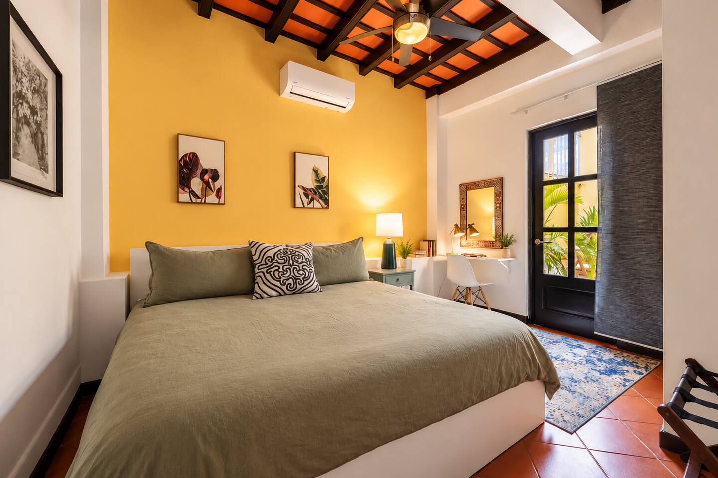 Relax in this inviting bedroom featuring a comfortable bed, warm Caribbean colors, exposed wood-beam ceilings, quiet air conditioning, & a ceiling fan. A dedicated workspace & direct access to the charming interior patio add comfort and convenience.